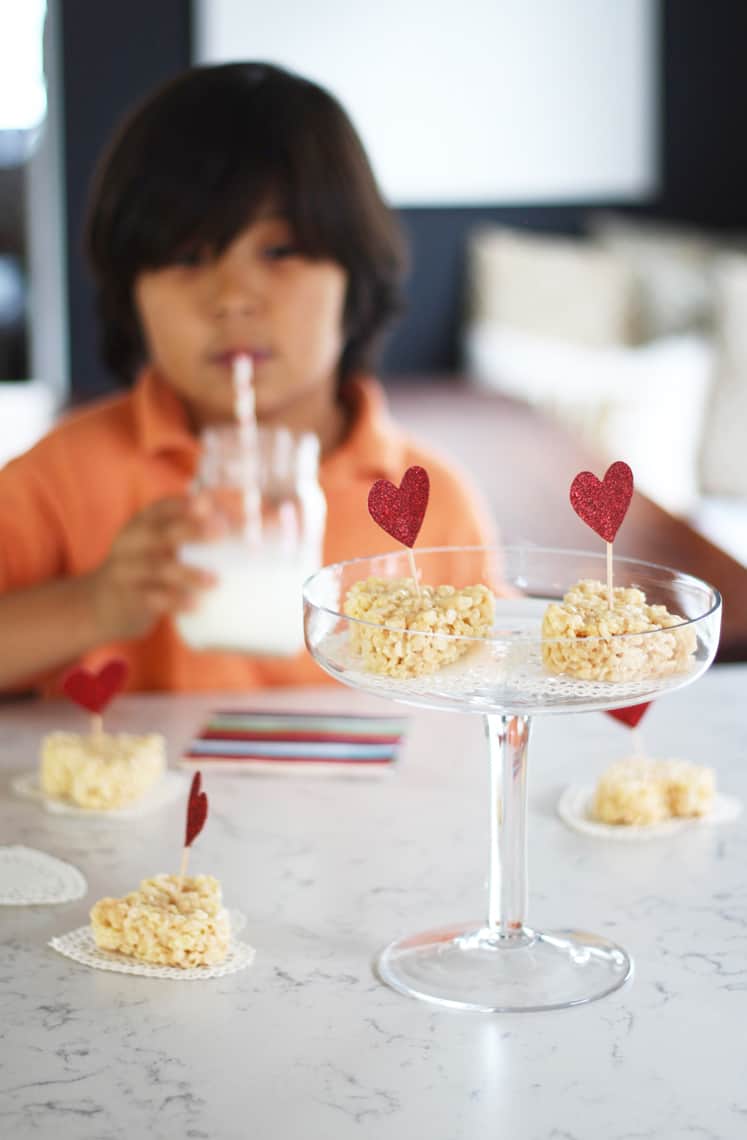 rice krispie valentine heart treats on a white countertop with a glass of milk with child